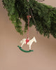 A Christmas tree branch displays a delightful Maileg Metal Ornament, Rocking Horse, featuring a white rocking horse with a red saddle and delicate hand-painted details, suspended by a red and white string against a neutral backdrop.