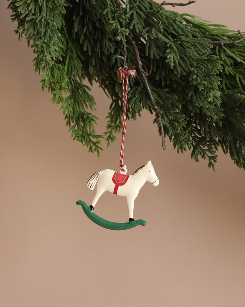 A Christmas tree branch displays a delightful Maileg Metal Ornament, Rocking Horse, featuring a white rocking horse with a red saddle and delicate hand-painted details, suspended by a red and white string against a neutral backdrop.