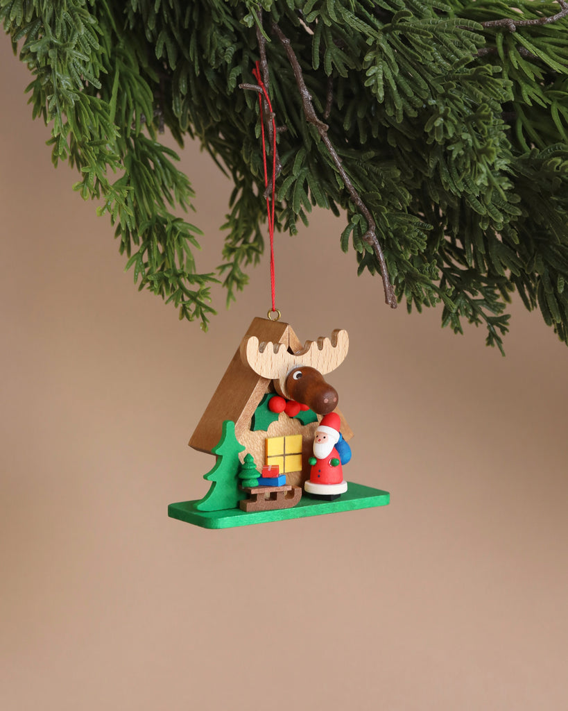 A festive Christian Ulbricht ornament, named "Santa with Elk House," hangs from a green tree branch. The piece features a moose with a prominent brown nose, accompanied by a small Santa figure, a yellow gift, and a green tree on its flat green base. Made in Germany, it stands out beautifully against the plain beige background.