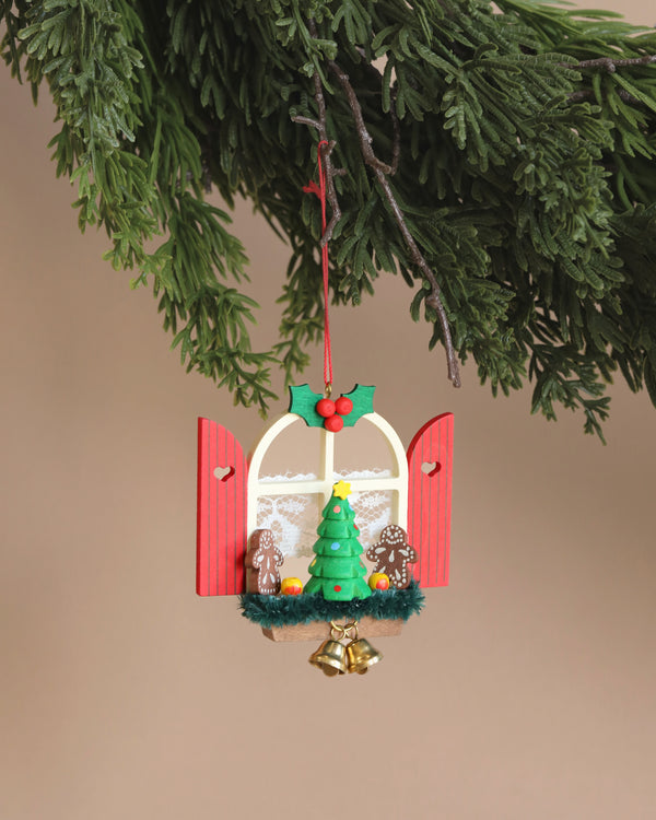 A Christian Ulbricht Ornament - Window with Gingerbread hangs from a pine branch. Hand-painted and made in Germany, it showcases a window with red shutters, a small Christmas tree, gingerbread figures, and gold bells. It’s beautifully adorned with holly leaves and berries on top.