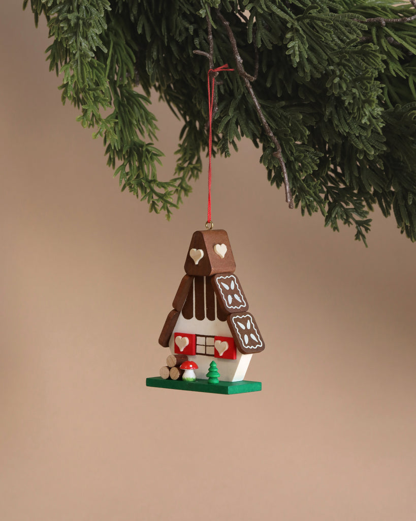 The Christian Ulbricht Ornament - Gingerbread House elegantly hangs from a branch against a neutral setting, adorned with heart and icing decorations. Expertly crafted in Germany, it boasts small red mushrooms and a green tree at its base. Please be aware that small parts may pose a choking hazard.