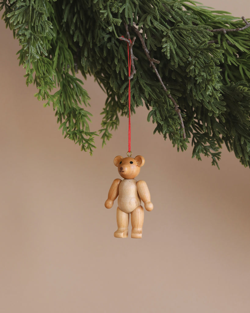 The Christian Ulbricht Ornament - Teddy Bear, a small wooden figurine, hangs from a red string on a green evergreen branch against a beige background.