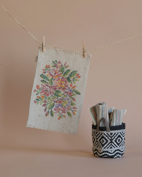 The Maileg Flower Rug, Medium, featuring a floral embroidery on beige fabric, hangs gracefully from a clothesline with wooden pegs. Next to it sits a charming black and white patterned miniature basket, reminiscent of those in cozy dollhouses, cradling folded striped fabrics. The backdrop is a simple yet warm beige hue.