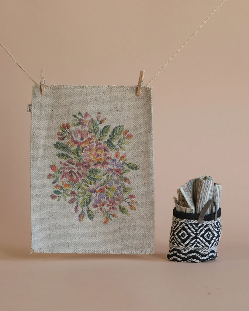 A Maileg Christmas Set featuring a floral tapestry adorned with colorful flowers is hung on a line with clothespins. Next to it, a black-and-white woven basket holds folded textiles. The neutral beige background adds warmth to this cozy corner; however, please keep small parts away from children under three due to potential choking hazards.