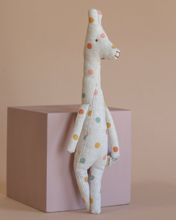 A tall, plush Maileg Mini Giraffe Stuffed Animal from the Safari Friend collection is resting against a mauve cube. It boasts a minimalist design with pastel spots on a beige background. The plain, light brown backdrop enhances its playful charm in this setting.