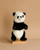 A small Panda Puppet stands on a light wooden base against a plain beige background. The toy, part of a range of realistic plush toys, features a classic panda color pattern with black arms, legs, ears, and eye patches, complemented by white fur on the rest of its body.