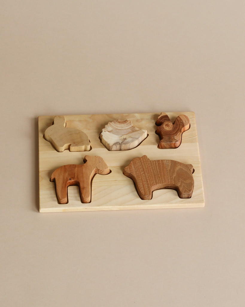 A Handmade Wooden Woodland Puzzle featuring five animal-shaped pieces in various shades of wood. The animals include a bear, a sheep, a rooster, a rabbit, and a seashell. Each piece is finished with linseed oil and beeswax for added durability and beauty. The board and pieces are set against a plain beige background.