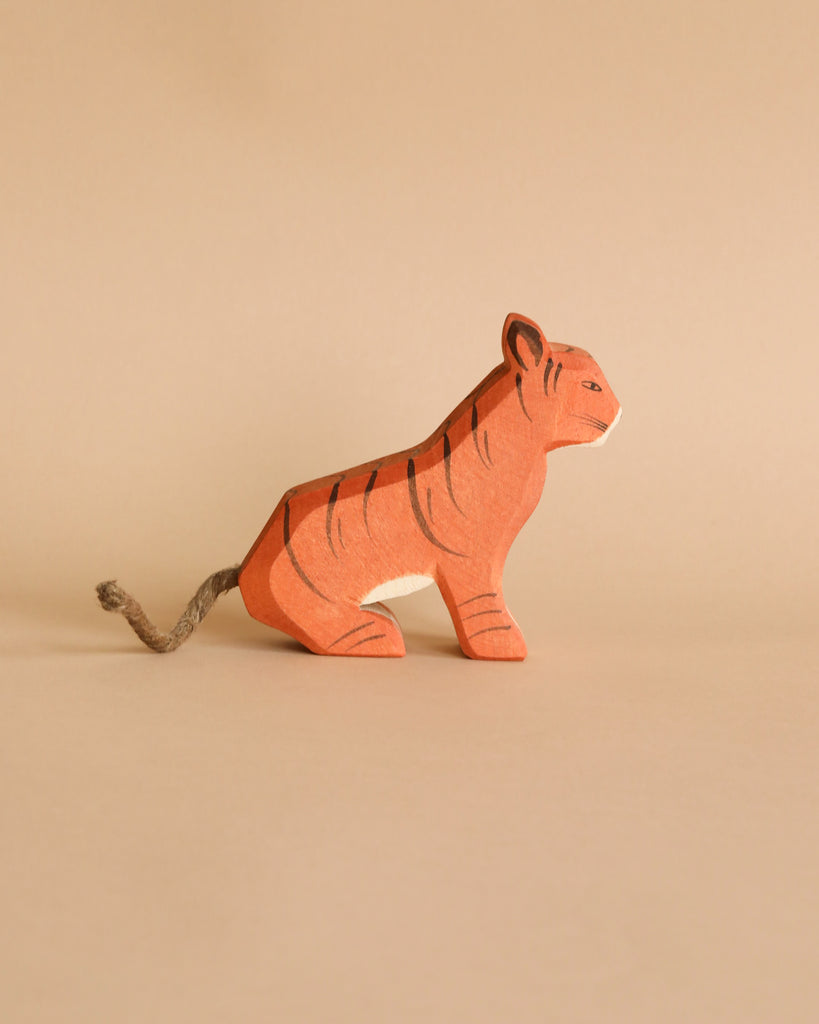 A small handcrafted wooden figurine of an orange tiger with black stripes and a white belly is depicted against a beige background. The tiger sits on its hind legs with its tail extended behind. The minimalist detailing and handmade appearance evoke the charm of the Ostheimer Sitting Tiger, perfect for imaginative play.