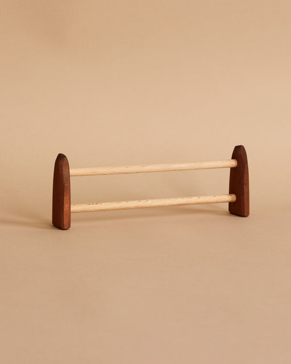 A minimalist wooden rack with two light-colored horizontal bars and two darker-colored, rounded triangular end supports, reminiscent of the quality found in handcrafted wooden toys, set against a beige background could be described as an Ostheimer Large Fence.