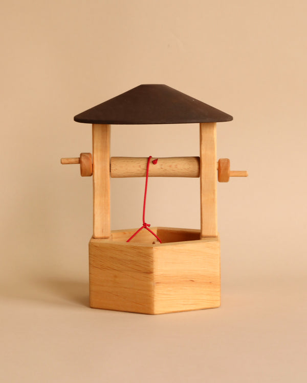 A small Ostheimer Wishing Well with a hexagonal base, a pitched roof, and a handle with a red string and bucket attached invites imaginative play. The handcrafted toy's simple and minimalist structure is set against a plain beige background, reminiscent of Ostheimer wooden toys.