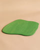 A green, irregularly-shaped Ostheimer Large Meadow Plate placed on a beige surface. The plate has a smooth texture and a natural wood grain pattern visible, reminiscent of Ostheimer toys and perfect for imaginative play.