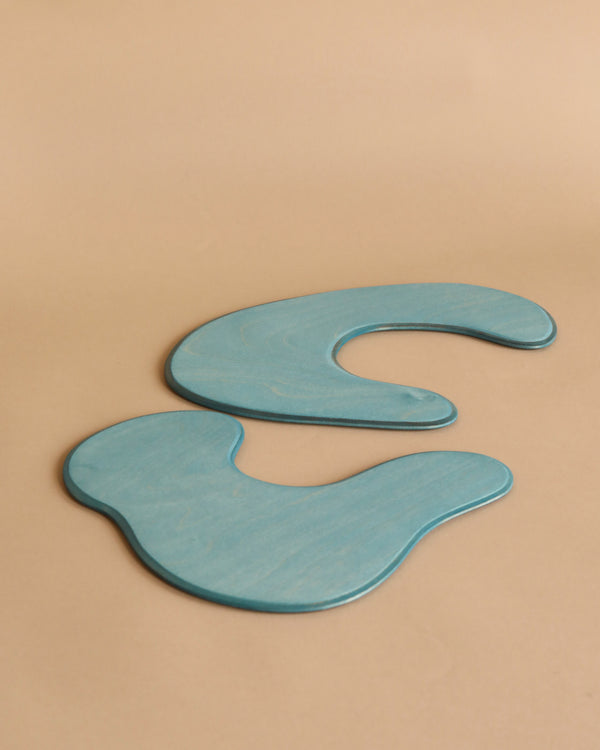 Two Ostheimer Pond Plates - Set of 2, abstract, curvilinear wooden shapes painted in light blue are placed on a beige surface. Their flowing, organic forms give a sense of fluidity and artistic design, reminiscent of handcrafted wooden toys. The smooth, polished shapes encourage imaginative play while contrasting subtly with the neutral background.