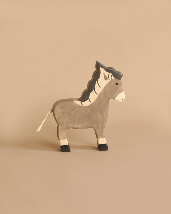 A simple Holztiger Small Donkey with a light beige body, white mane, and black hooves is standing on a plain, beige background. This handcrafted wood toy features minimalistic painted details and has a short, white string tail. Made in Europe by HOLZTIGER figures.