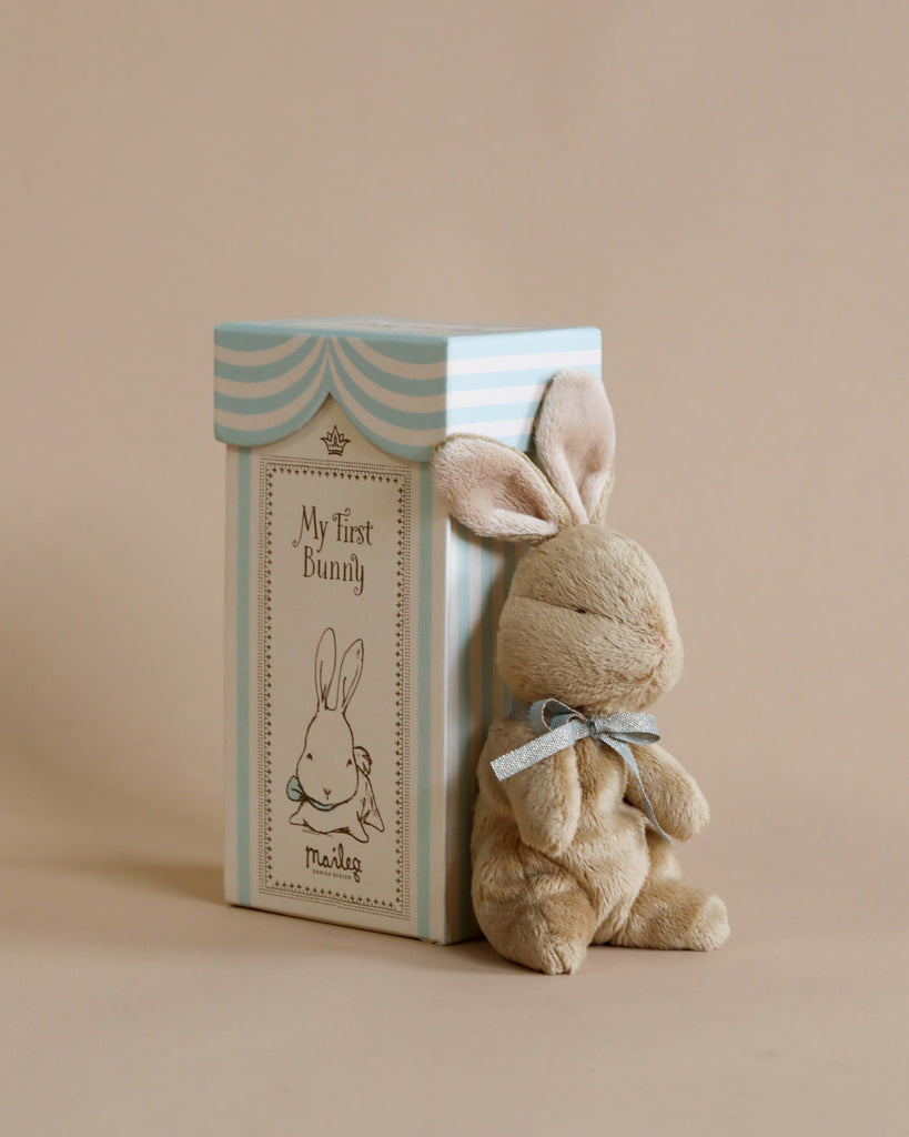 A soft, beige bunny plush toy with a blue ribbon around its neck sits next to a pastel blue and white box. The box is labeled "Maileg My First Bunny - Light Blue" and features an illustration of a bunny. This soft plush bunny makes the perfect baby shower gift against the neutral beige background.