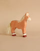 A small, wooden toy horse stands on a beige surface. Handcrafted with care, this light brown horse with a white mane and tail features red and white accents on its lower legs. The tail appears textured and is made from a different material. This Holztiger Horse, Standing, Light Brown figure is proudly made in Europe.