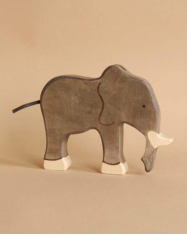 A wooden toy elephant stands on a neutral brown background. The handcrafted wood toy is painted in shades of grey with white tusks and feet, featuring simple, minimalist details. This delightful piece is the Holztiger Elephant, part of the renowned HOLZTIGER figures collection, proudly made in Europe.