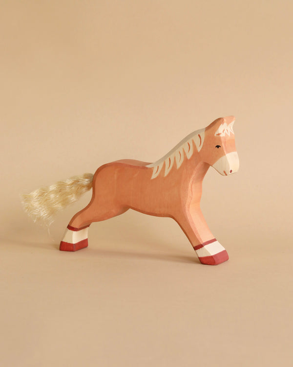 A Holztiger Horse, Running, Light Brown made from maple and beech wood features a light brown body, off-white mane, and tail. It stands on a beige background, with two red-tipped hooves and a painted white stripe on its legs. The horse has a simple yet handcrafted appearance akin to HOLZTIGER figures with minimalistic features.