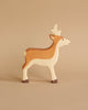 A handcrafted wooden toy roe buck stands on a beige surface against a matching beige background. The roe buck, part of the renowned Holztiger Roe Buck figures collection, is painted in natural shades of brown and white, with dark brown at the tips of its hooves and light brown shading on its maple and beech wood body.