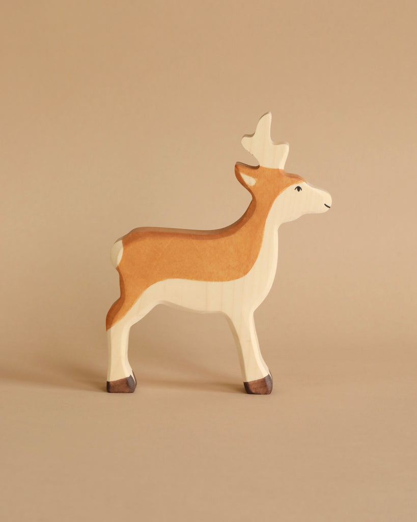 A handcrafted wooden toy roe buck stands on a beige surface against a matching beige background. The roe buck, part of the renowned Holztiger Roe Buck figures collection, is painted in natural shades of brown and white, with dark brown at the tips of its hooves and light brown shading on its maple and beech wood body.