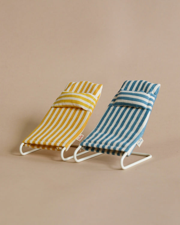 Two Maileg Beach Chairs And/Or Umbrella with white metal frames. One chair has yellow and white stripes, and the other has blue and white stripes. Both chairs have a matching small pillow at the top. The background is a plain beige color.