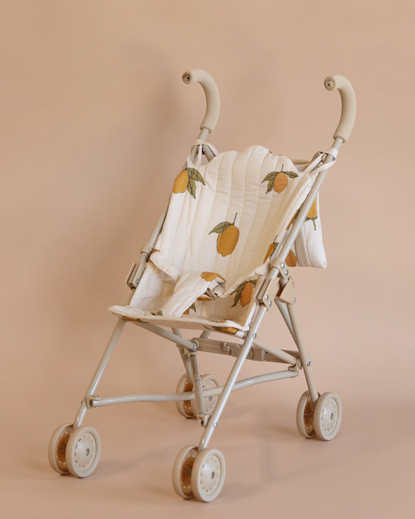 The Konges Sloejd Doll Stroller - Grand Lemon features a beige frame, removable lemon-printed fabric, and double wheels. It is styled with charming curved handles and is displayed on a warm, earth-toned background.