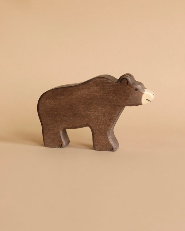 A wooden toy bear stands on a neutral background. The Holztiger Brown Bear figure is simplified in design and has a natural brown finish with lighter details on its face and nose. The overall aesthetic is minimalistic, resembling painted watercolours, highlighting its handcrafted wood charm.