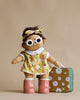 A Dinkum Doll - Sprout (Extended Pack) with brown hair wearing white round sunglasses, a floral headband, and a matching floral dress with a white collar. The soft cotton doll has beige skin tone and wears pink boots. It boasts embroidered details and stands next to a small green and mustard-colored suitcase with flower patterns.