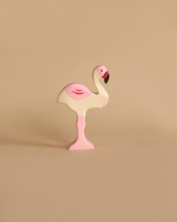 A small, pastel-colored wooden flamingo toy stands on one leg against a beige background. Carefully handcrafted from high-quality wood, this Holztiger Flamingo is painted in light shades of pink and white, with a slightly darker pink on its wing and a black beak.