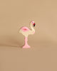 A small, pastel-colored wooden flamingo toy stands on one leg against a beige background. Carefully handcrafted from high-quality wood, this Holztiger Flamingo is painted in light shades of pink and white, with a slightly darker pink on its wing and a black beak.