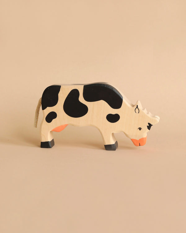 A Holztiger Grazing Black Cow with a natural finish and black spots stands against a beige background. Handcrafted in Europe by HOLZTIGER figures, the cow features an orange snout and black hooves, exuding charm and quality.