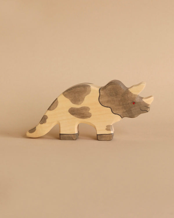 A handcrafted wood triceratops stands against a plain, beige background. The toy features a light wood finish with dark, painted spots and a small red eye. Its silhouette highlights three horns and a frilled neck typical of Holztiger Triceratops figures, ensuring quality Made in Europe craftsmanship.