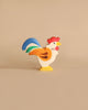 A Holztiger Rooster, made in Europe and painted in bright colors, features a red comb, yellow beak, white body with orange feathers, and a blue and green tail. This handcrafted wood piece stands upright on a neutral tan background and epitomizes the charm of HOLZTIGER figures.