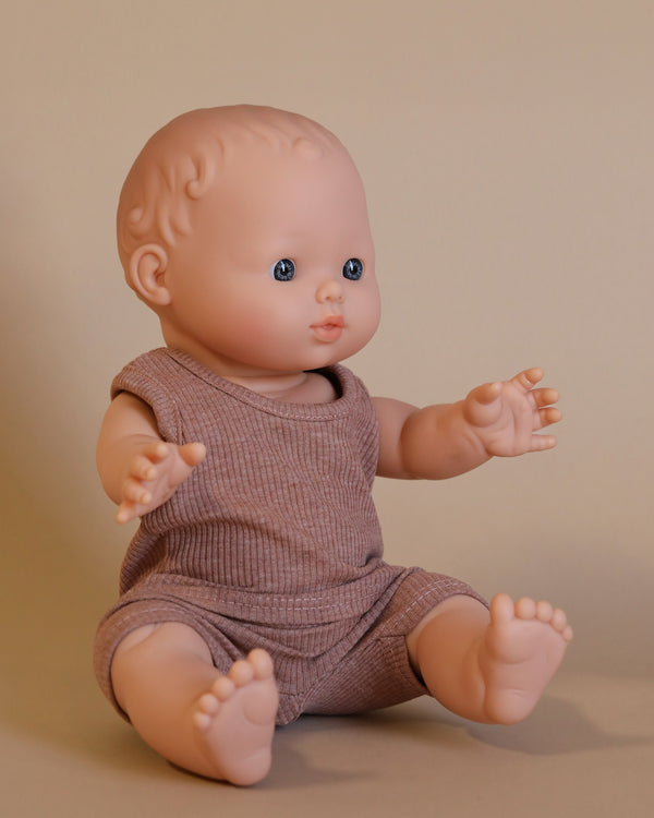 A lifelike Minikane Doll (13") - César, dressed in a ribbed brown outfit, is seated against a plain beige background. Made from phthalate-free vinyl for safe imaginative play, the doll showcases detailed blue eyes and slightly curled hair, with its arms and legs positioned outward.