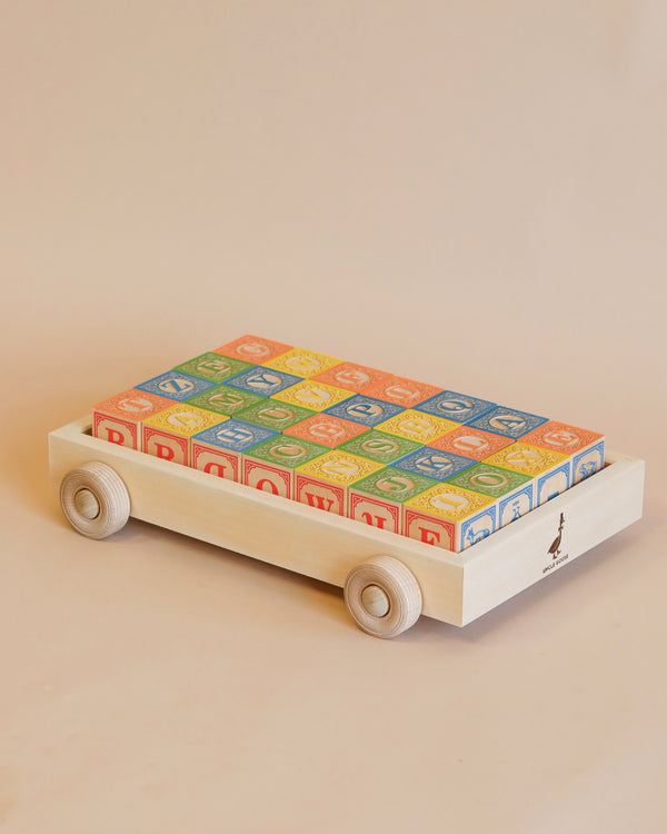 A handmade basswood wagon with four wheels carries the Uncle Goose Classic ABC Blocks with Wagon. The blocks feature letters, numbers, and decorative designs in blue, red, yellow, and green. The charming cart and its vibrant contents rest against a beige background.