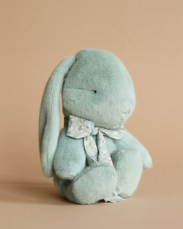 A Maileg Small Plush Bunny - Mint with long ears and a floral bow around its neck, sitting against a plain beige background. Its eyes appear closed and its overall color is soft blue.