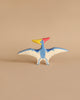 A small, colorful wooden toy figurine of a pteranodon with blue wings, a red crest, a yellow beak, and white underwings decorated with blue dots, stands against a plain beige background. Handcrafted out of maple and beech wood, this Holztiger Pteranodon Dinosaur is made in Europe.