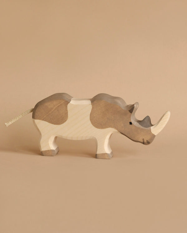 A wooden toy rhinoceros stands on a beige background. The Holztiger Rhinoceros is painted in shades of gray and white, with a small black eye and a tail made of string. Its two horns are prominently carved on its head, showcasing the handcrafted detail typical of wooden toys.