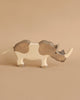 A wooden toy rhinoceros stands on a beige background. The Holztiger Rhinoceros is painted in shades of gray and white, with a small black eye and a tail made of string. Its two horns are prominently carved on its head, showcasing the handcrafted detail typical of wooden toys.