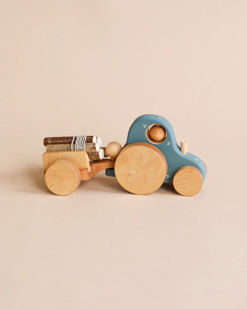 A Handmade Wooden Tractor with a blue cab and detachable natural wood trailer carries small logs secured by a string. The toy has simple, rounded shapes and is placed on a plain beige background.