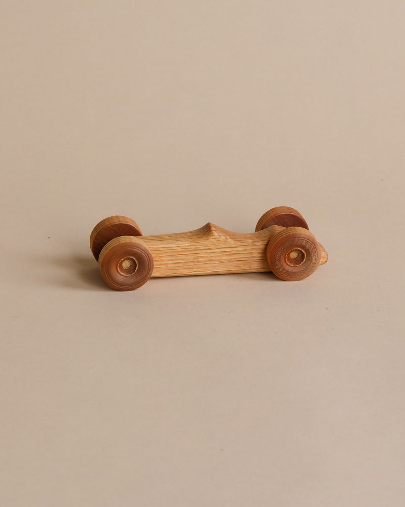 A Handmade Wooden Race Car with four round wheels and a smooth, minimalist design sits on a beige surface. The classic car has an oak wood finish, showcasing the natural grain and texture of the material.