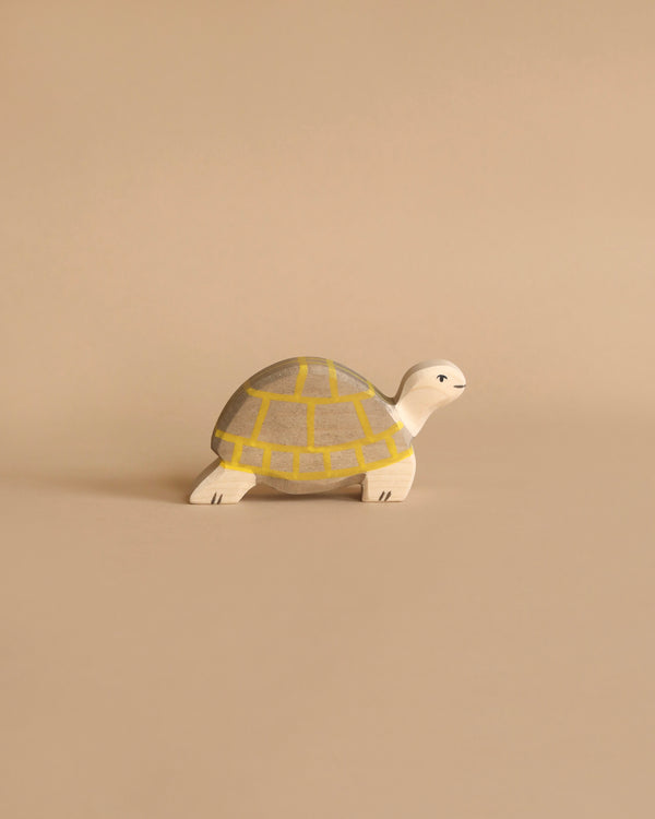 A small, handcrafted wooden toy turtle placed on a beige background. The Holztiger Tortoise, reminiscent of classic HOLZTIGER figures, has a white head and legs with a soft grey shell accented by yellow grid patterns. It is carefully crafted from maple and beech wood.