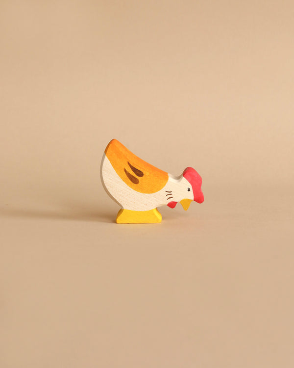 A small, colorful wooden figurine of a hen stands on a plain beige background. Handcrafted out of maple and beech wood, this Holztiger Hen, Pecking figure is painted with yellow, white, and orange body parts and has a red comb on its head. The simple, cartoonish design adds a playful touch to the figurine.