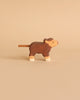 A small wooden toy cow with a rope tail is depicted against a beige background. Made in Europe, the Holztiger Highland Cattle, Small figure is brown with white accents on its legs and face, featuring a simple, hand-painted design.