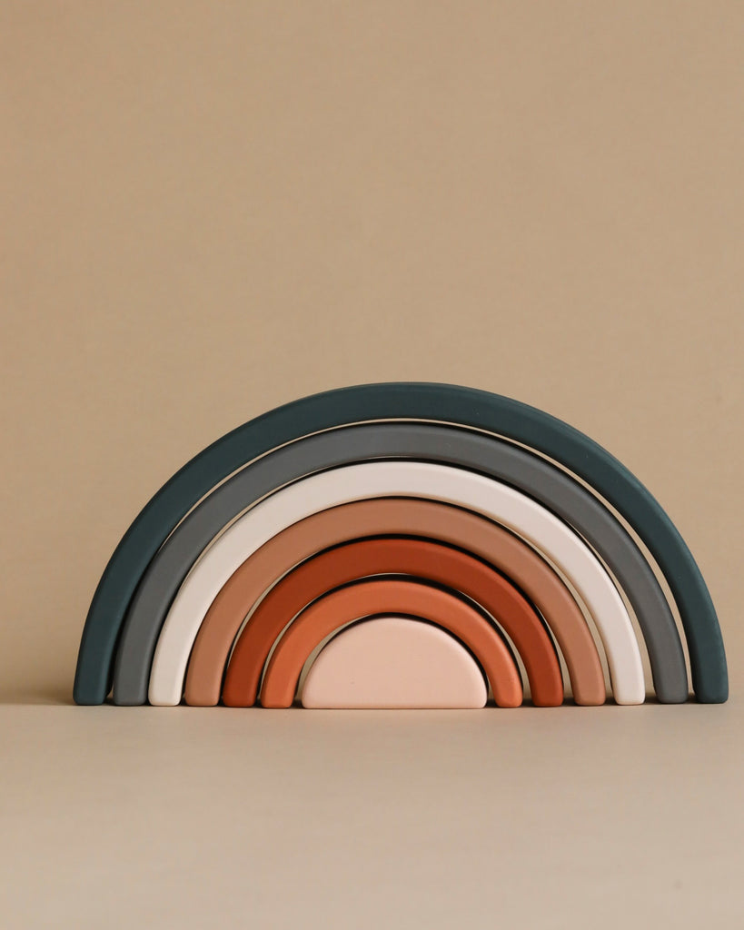 The Handmade Rainbow Stacker - Mojave Desert is a minimalistic, handcrafted toy featuring seven curved, arc-shaped pieces in muted colors like green, beige, white, terracotta, and peach. Each piece is painted with non-toxic paint and arranged by size from largest to smallest on a neutral background.