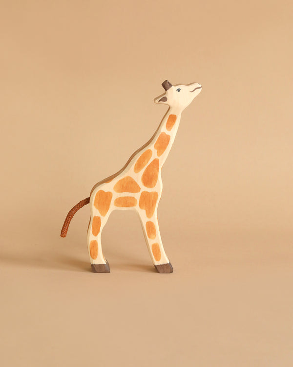 A Holztiger Small Feeding Giraffe, made in Europe, stands against a beige background, featuring light brown spots, a brown tail, dark brown hooves, and a small pair of brown horns on its head. The giraffe's head is lifted as if looking upwards. This piece is reminiscent of HOLZTIGER figures.