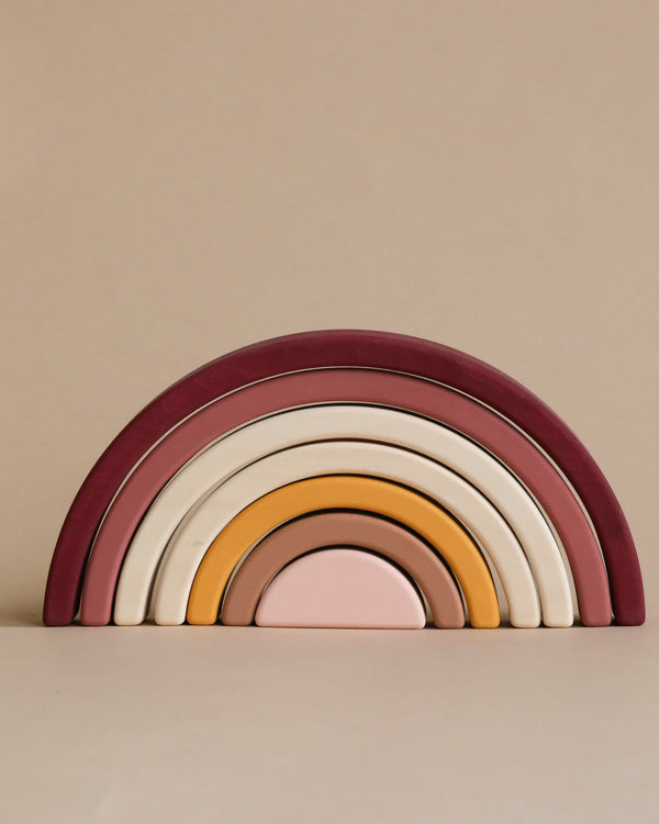 The Handmade Rainbow Stacker - Pink is a minimalist wooden toy featuring nested arches painted in maroon, pink, white, yellow, and purple. Each piece is lovingly hand-painted with non-toxic paint. It’s displayed against a plain beige wall.