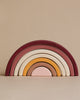 The Handmade Rainbow Stacker - Pink is a minimalist wooden toy featuring nested arches painted in maroon, pink, white, yellow, and purple. Each piece is lovingly hand-painted with non-toxic paint. It’s displayed against a plain beige wall.