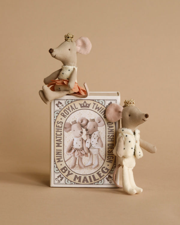 Two small, Royal Twins in Box fabric mouse figurines with crowns, one seated on a book and one standing beside it, against a beige background. The book features intricate illustrations of mice. One mouse is dressed in