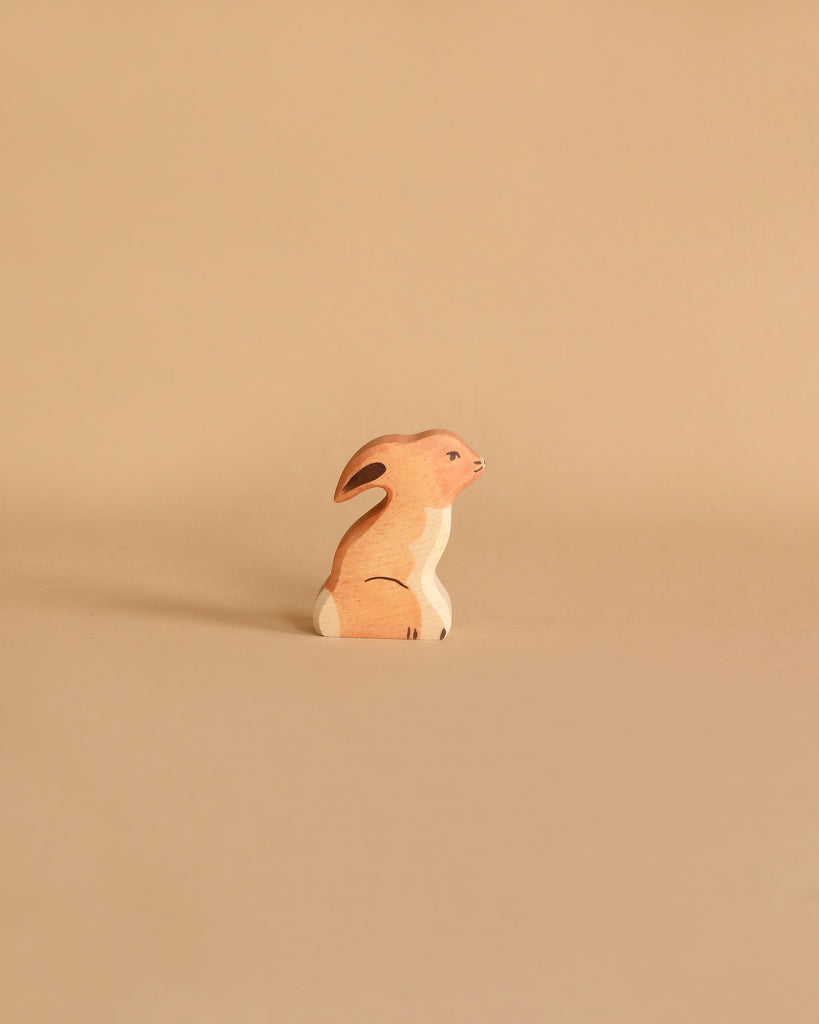 A small wooden figurine of a rabbit, portrayed in a sitting position, is placed against a neutral beige background. This Holztiger Rabbit, Sitting is light brown with white accents on its chest and tail. Made in Europe like all HOLZTIGER figures, it exudes quality and charm.