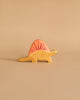 A Holztiger Dimetrodon Dinosaur with a yellow body and a pink, fin-like structure on its back stands against a light beige background. This handcrafted wood figure features a simple design, painted eyes, and minimal detailing, reminiscent of the charming HOLZTIGER figures made in Europe.